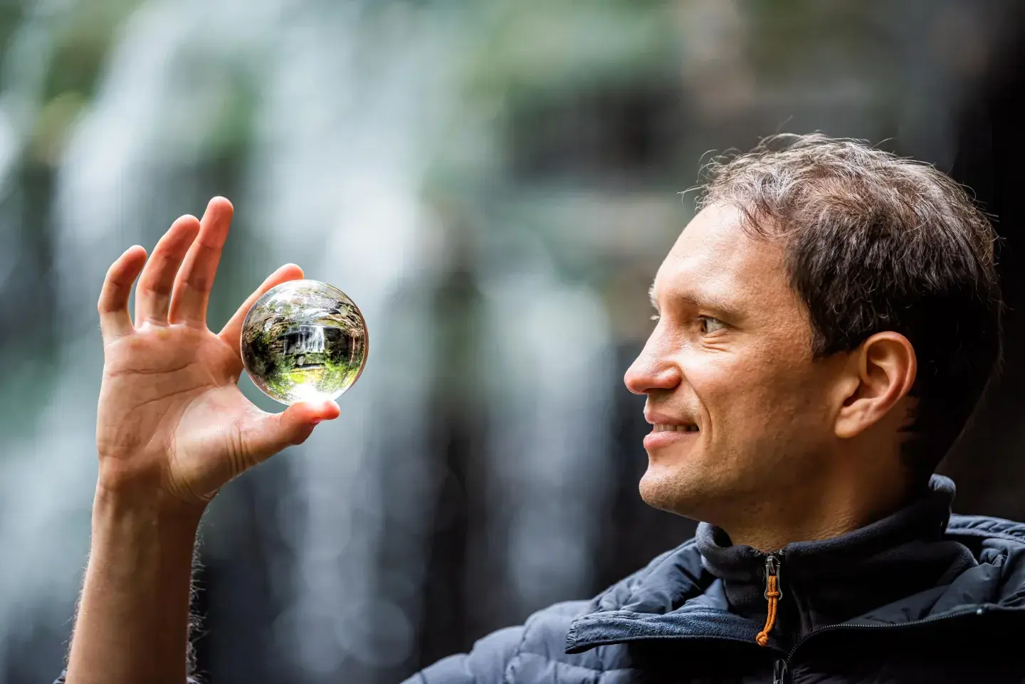 An individual holding a clear glass orb that reflects a forest scene, with a waterfall and greenery softly out of focus behind them.