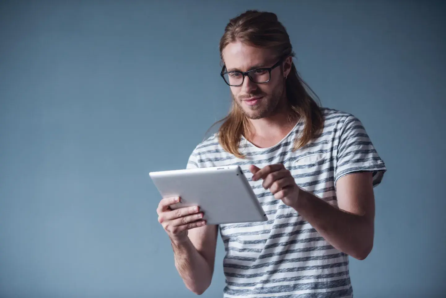 “A person standing against a plain blue background, holding a tablet with one hand and using the other to interact with the screen.