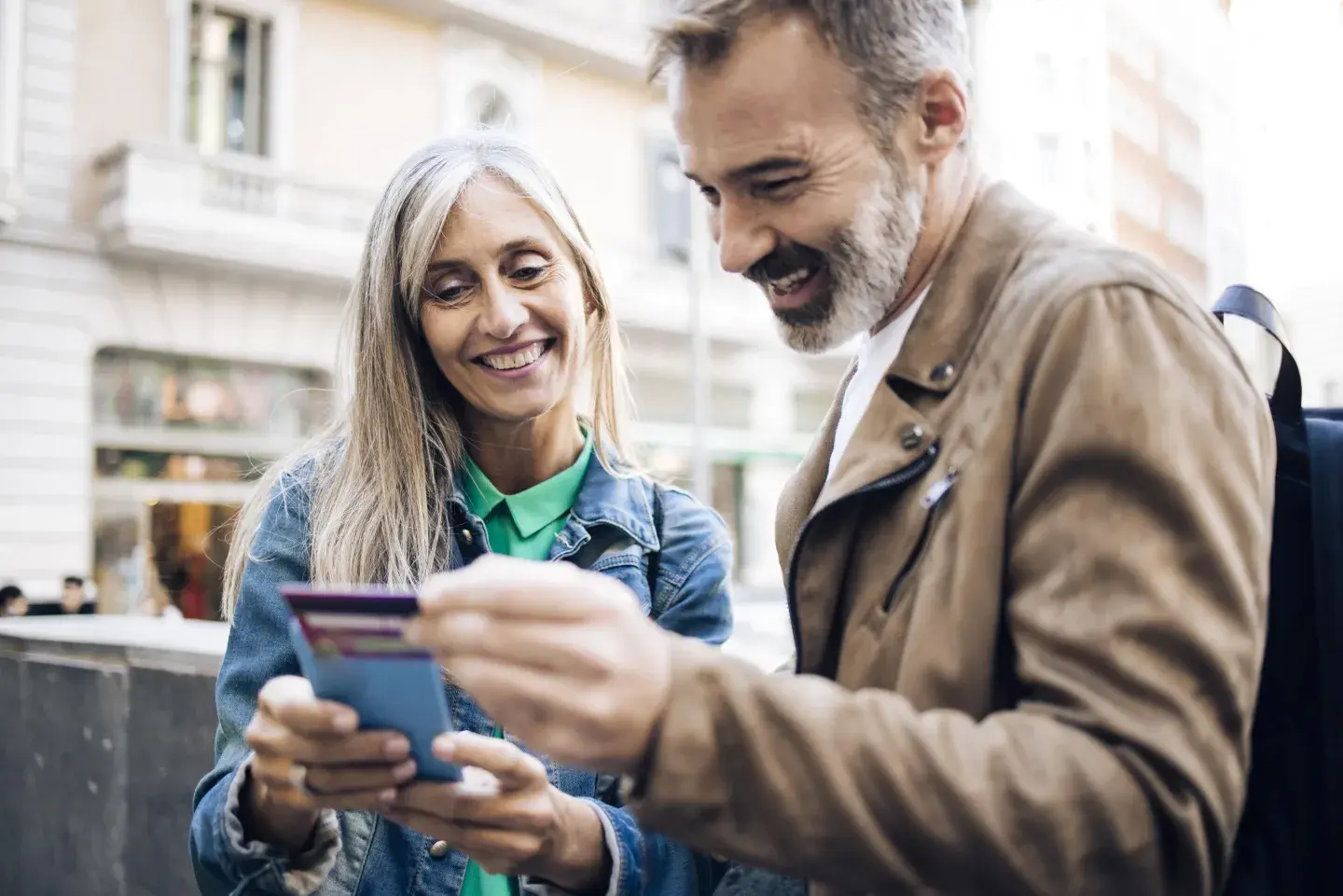 Two people review a card and a mobile phone while standing outside near buildings in an urban setting.