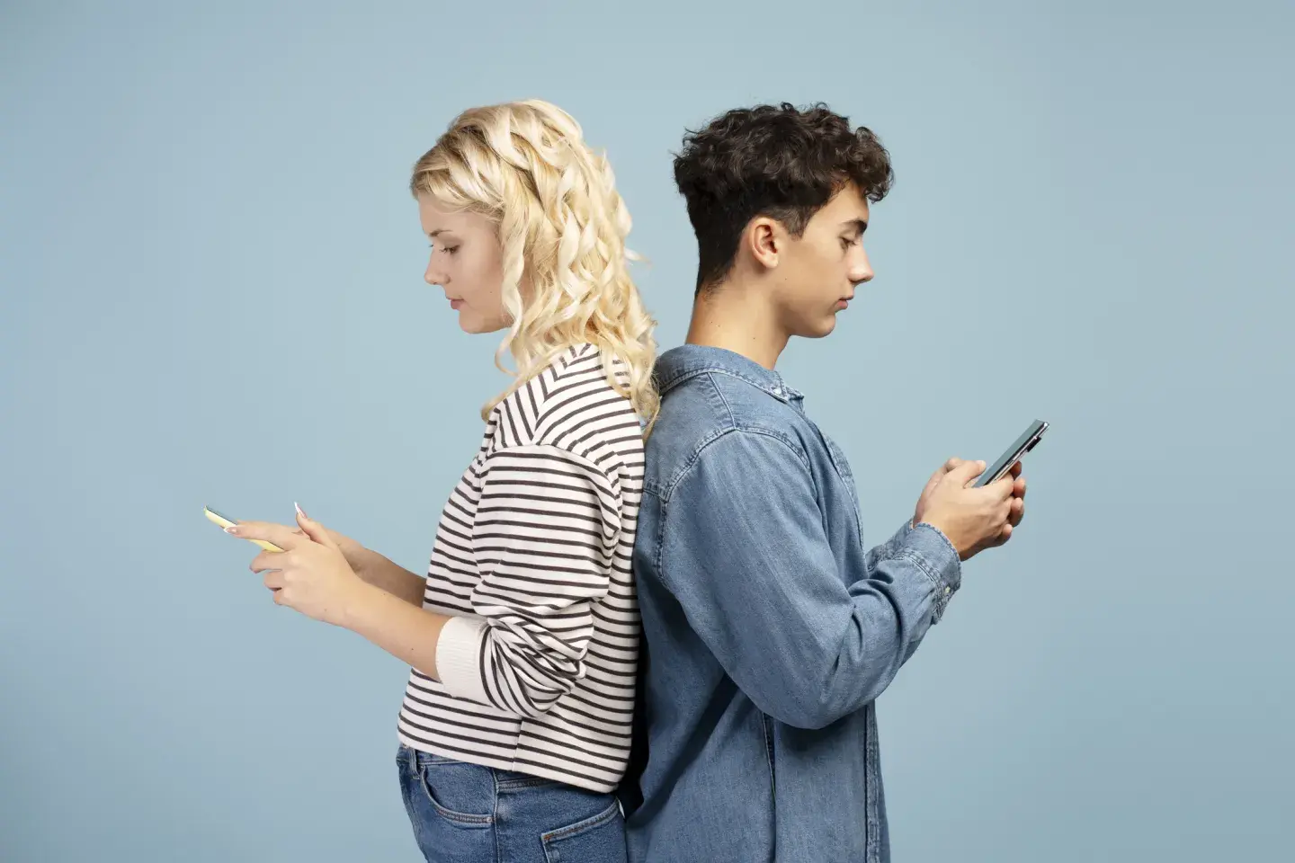 Two people standing back to back against a solid blue background, each holding and looking at a smartphone